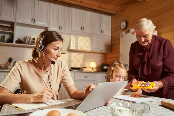 Responsible female remote worker making notes in notebook during video conference while grandmother showing new toy to girl